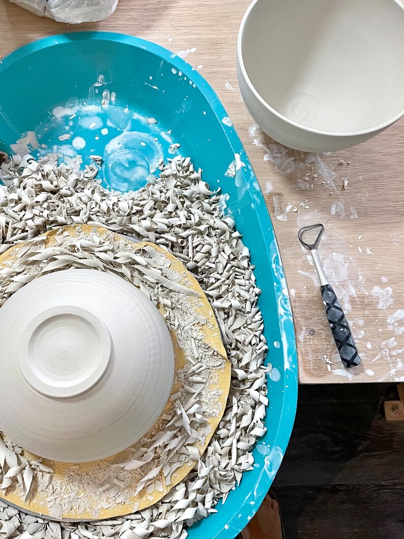 May include: A white ceramic bowl sits on a pottery wheel, surrounded by white clay shavings. A turquoise plastic tray holds the wheel and shavings. A second white ceramic bowl sits on a wooden table next to the tray.