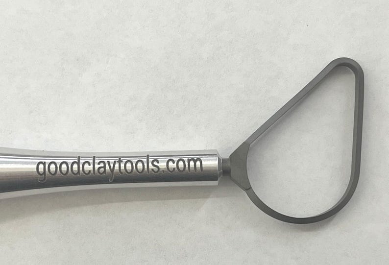 May include: A silver sculpting tool with a triangular loop at the end and a handle with the text "goodclaytools.com" engraved on it.