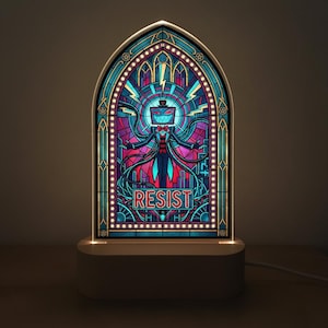 Può includere: Lampada a LED in stile vetrata raffigurante una figura con testa a schermo TV e la scritta "RESIST". La lampada ha una base bianca e una luce calda, con una forma ad arco gotico. Il design utilizza colori vivaci.