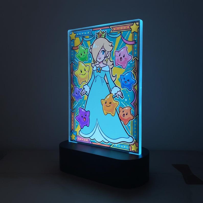 Op de afbeelding: Een rechthoekige LED-lamp met een cartoonillustratie van een personage in een blauwe jurk en kroon, omringd door kleurrijke sterren. De lamp heeft een zwarte basis en een blauw verlichte rand, wat een levendige weergave cre&euml;ert.