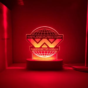 Op de afbeelding: Een rood LED-licht met een witte basis heeft het Weyland-Yutani Corp.-logo. Het logo is een gestileerde "W" met een globe erachter. De tekst "Building Better Worlds" staat onder het logo.