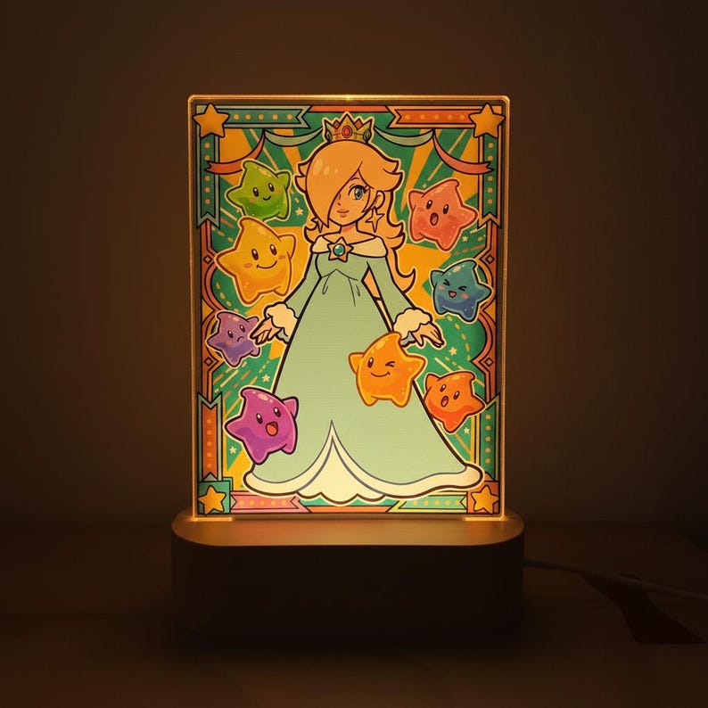 Op de afbeelding: Een verlicht acryl paneel met een cartoon illustratie van een personage in een teal jurk en kroon, omringd door kleurrijke sterren. Het paneel staat op een donkere basis en straalt een warme gloed uit.