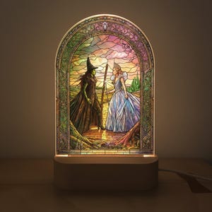 Puede incluir: Lámpara LED estilo vidriera con una escena de El Mago de Oz. La obra representa a la Malvada Bruja del Oeste y Glinda la Bruja Buena. La lámpara tiene una base blanca y un diseño arqueado colorido con un borde verde y dorado.