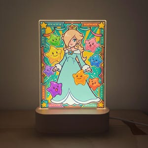 Op de afbeelding: Een verlicht acryl paneel met een cartoonprinses in een blauwe jurk, omringd door kleurrijke sterren. Het paneel staat op een witte basis met een netsnoer, waardoor een decoratief nachtlampje ontstaat.