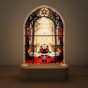Könnte beinhalten: Eine Lampe im Buntglasstil mit einem Cartoon-Lammcharakter mit roten Augen und einem schwarz-weißen Design. Die Lampe hat einen Holzsockel und zeigt eine Waldszene mit Kerzen und einem Pentagramm.