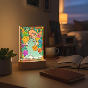 Op de afbeelding: Decoratief nachtlampje met een stripfiguur in een turquoise jurk, omringd door kleurrijke sterren. Het licht staat op een houten voet en is verlicht, waardoor een warme gloed ontstaat. Het ontwerp doet denken aan glas-in-loodkunst.