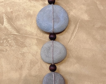 Puede incluir: Una pieza de arte colgante de piedra con cuatro piedras grises de forma ovalada conectadas por una fina cuerda de color claro. Cuentas de madera marrón oscuro separan las piedras. Las piedras son de diferentes tamaños.