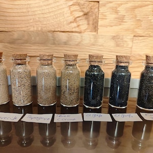 May include: Seven clear glass bottles with cork stoppers, each filled with different coloured sand ranging from light beige to dark black. Each bottle is placed behind a small white paper label with handwritten text identifying the sand source.