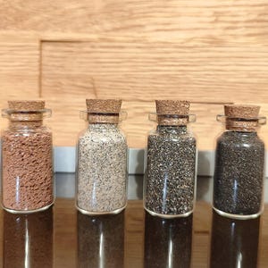Puede incluir: Cuatro botellas pequeñas de vidrio transparente con tapones de corcho, cada una llena con una sustancia granular de diferente color. El contenido varía desde un marrón rojizo, beige claro, gris oscuro hasta negro sólido.