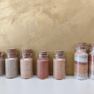 Puede incluir: Una colección de pequeños frascos de vidrio llenos de arenas de varios colores. Cada frasco tiene un tapón de corcho. Las arenas varían en color de blanco a marrón rojizo. Los frascos están colocados sobre una superficie blanca.