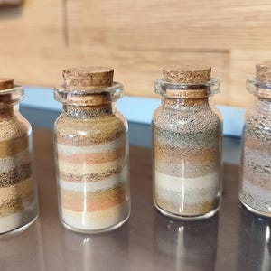 Könnte beinhalten: Vier kleine Glasflaschen mit Korken, jede gefüllt mit Schichten aus unterschiedlich farbigem Sand. Die Sandfarben umfassen Weiß, Beige, Braun und Orange, wodurch ein gestreifter Effekt entsteht. Die Flaschen werden auf einer dunklen Oberfläche ausgestellt.