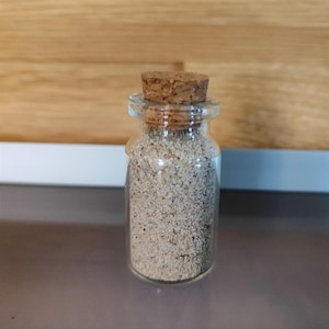 Puede incluir: Un pequeño frasco de vidrio con tapa de corcho lleno de una sustancia arenosa de color marrón claro.
