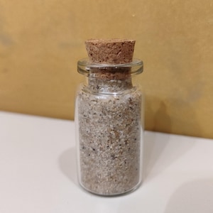 Könnte beinhalten: Eine kleine Glasflasche mit einem Korkverschluss, gefüllt mit Sand. Die Flasche ist klar und der Sand ist hellbeige.