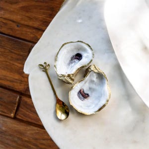 May include: Two oyster shells with gold-painted edges and a small gold spoon with a leaf design. The shells have a pearlescent interior and are arranged on a white marble surface. The spoon is next to the shells.