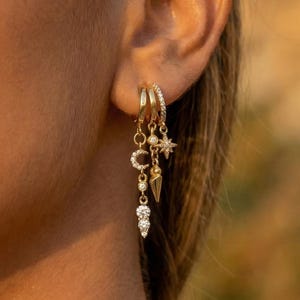 May include: Gold hoop earrings with dangling charms. The earrings feature a crescent moon, star, and teardrop-shaped charms, along with a dangling cluster of clear crystals. The earrings are worn on a person's ear, with a blurred background.