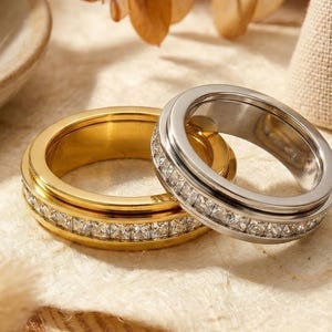 Könnte beinhalten: Zwei Ringe, einer goldfarben und einer silberfarben, werden gezeigt. Der goldfarbene Ring hat ein Band aus kleinen, klaren Steinen. Der silberfarbene Ring hat ebenfalls ein Band aus klaren Steinen. Beide Ringe haben ein schlichtes, elegantes Design.