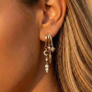 以下が含まれることがあります： ゴールドトーンのフープピアス。三日月、星、しずく型のチャームと、複数の透明で輝く石が特徴です。ピアスは、金髪の人の耳に着用されています。