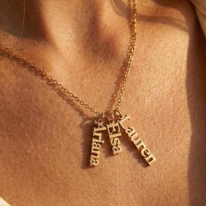 Könnte beinhalten: Eine goldfarbene Halskette mit drei vertikalen Anhängern mit den Namen "Ariana", "Elsa" und "Lauren". Die Namen sind auf den Anhängern eingraviert. Die Halskette wird um den Hals getragen.