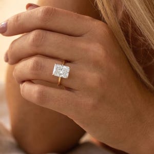 Könnte beinhalten: Ein goldener Ring mit einem großen, rechteckigen, klaren Edelstein. Der Ring befindet sich an einem Finger, mit einem goldenen Armband am Handgelenk. Der Hintergrund ist weich verschwommen, mit neutralen Tönen.