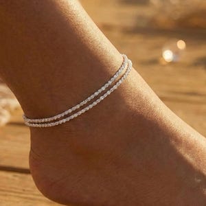 Puede incluir: Una delicada tobillera de doble hebra con un diseño brillante y con cuentas. La joya plateada se lleva en el tobillo, mostrando un accesorio simple pero elegante. La tobillera añade un toque de brillo al pie.
