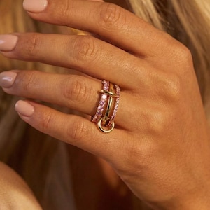 Könnte beinhalten: Ein goldener Ringsatz mit drei gestapelten Bändern. Ein Band ist mit rosa Edelsteinen besetzt, ein anderes ist schlicht goldfarben, und das dritte hat einen horizontalen Goldstab. Ein kleiner goldener Anhänger hängt unten.