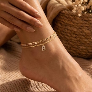 Peut inclure: Un bracelet de cheville doré avec deux chaînes, l'une avec un motif de maillons gourmette et l'autre avec une chaîne perlée délicate. Un petit charme avec la lettre "B" est suspendu à la chaîne inférieure. Le bracelet est porté sur un pied.