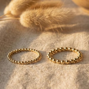 May include: Two gold beaded rings are displayed on a beige fabric surface. The rings are made of small, round beads, creating a textured appearance. The rings are in focus, with dried plants in the background.