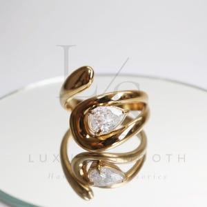 May include: A gold-colored ring with a pear-shaped, clear gemstone. The ring has a unique, swirling design, and is reflected in the mirrored surface below. The ring is a piece of jewelry.