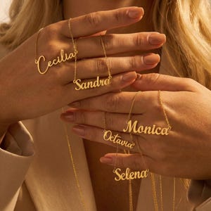 Könnte beinhalten: Goldfarbene Namensketten mit den Namen "Cecilia", "Sandra", "Monica", "Octavia" und "Selena". Die Halsketten werden vor einem neutralen Hintergrund präsentiert, der die zarte Schrift jedes Namens hervorhebt.