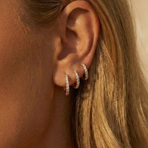 Puede incluir: Primer plano de tres pendientes de aro dorados, cada uno incrustado con pequeñas piedras preciosas brillantes. Los pendientes son de diferentes tamaños, creando un efecto en capas. La imagen muestra los pendientes sobre un fondo de cabello rubio y piel.