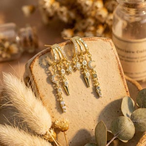 May include: Gold-toned hoop earrings with dangling charms. Each earring features a series of charms, including stars, moons, and teardrop shapes, all adorned with small, sparkling crystals. The earrings are displayed on a textured, light brown surface.