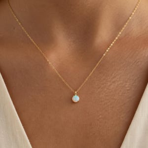 Op de afbeelding: Een delicate gouden ketting met een kleine, ronde opaal hanger. De opaal heeft een melkachtige witte kleur met subtiele iriserende flitsen. De ketting is fijn en de hanger is gecentreerd, wat een minimalistisch en elegant ontwerp creëert.