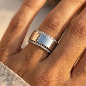 May include: A silver ring with a wide band and a rectangular center. The ring is accented with a row of small, sparkling stones along the top and bottom edges. The ring is on a finger.