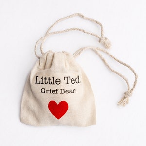 Puede incluir: Una peque&ntilde;a bolsa de tela beige con el texto "Little Ted. Grief Bear." impreso en la parte delantera. Un coraz&oacute;n rojo est&aacute; impreso debajo del texto.
