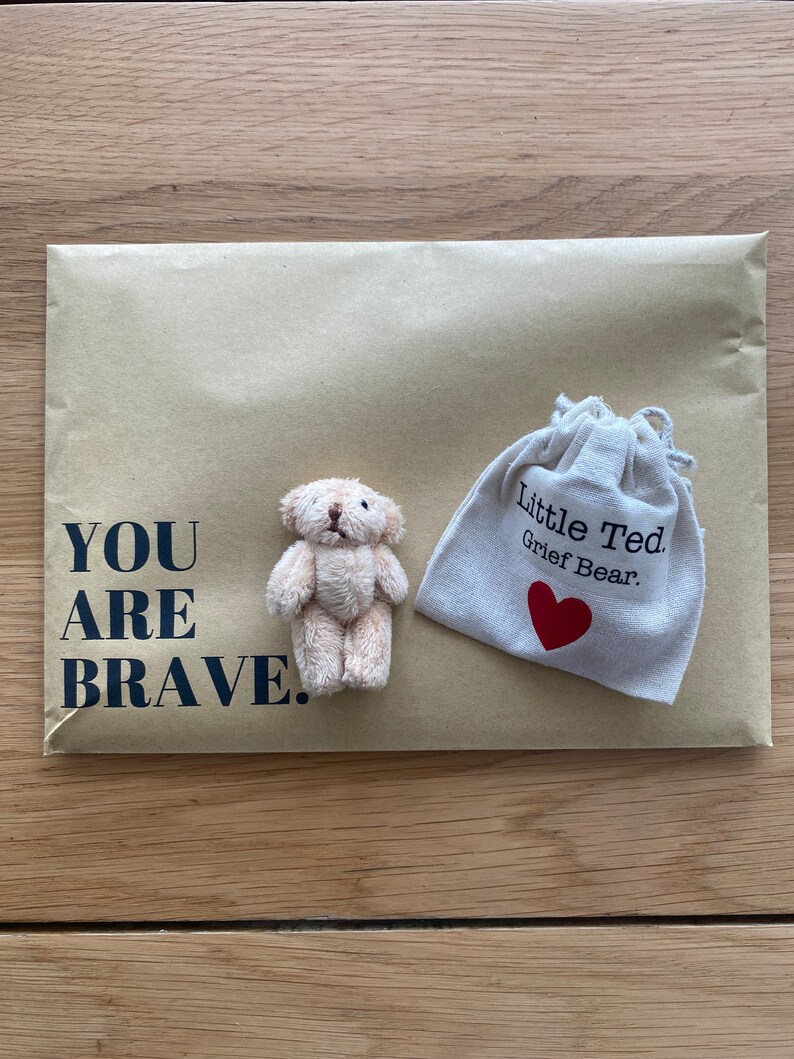 Puede incluir: Un peque&ntilde;o oso de peluche beige con un coraz&oacute;n rojo en un sobre de papel marr&oacute;n. El sobre tiene el texto "YOU ARE BRAVE." impreso en &eacute;l. Una peque&ntilde;a bolsa blanca con el texto "Little Ted Grief Bear." tambi&eacute;n est&aacute; en el sobre.