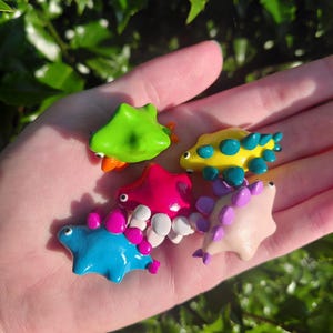 Op de afbeelding: Een verzameling van vijf kleine, gladde, handgebeeldhouwde dinosaurusfiguurtjes in felle kleuren, waaronder limoengroen, geel met groenblauwe stekels, felroze, blauw met magentakleurige stekels en lichtroze met paarse stekels, elk met kleine zwarte ogen.