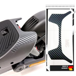 May include: A close-up of a black and gray carbon fiber textured scooter part. The image also shows a carbon fiber sticker with the text "MADE IN GERMANY". The scooter part has a black footrest and a yellow reflector.