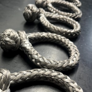 Könnte beinhalten: Nahaufnahme einer Reihe von silbernen geflochtenen Seilschlaufen mit Knoten an den Enden. Das Seil ist versetzt angeordnet, wobei sich jede Schlaufe über die darunterliegende überlappt.