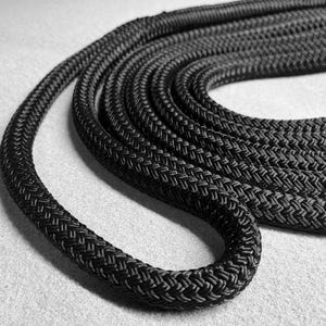 May include: Black braided rope coiled on a grey surface. The rope is thick and has a textured surface.