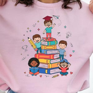 May include: A light pink crewneck sweatshirt featuring a colorful graphic print of five children interacting with a tall stack of books. The illustration includes a child wearing a graduation cap, a large yellow pencil, and small decorative icons like stars, hearts, and a paper airplane.