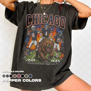 Può includere: Una t-shirt a maniche corte grigio antracite con una stampa grafica di tre giocatori di football scheletrici e un grande orso bruno su un campo. Il testo CHICAGO è stampato in grandi lettere arancioni e blu sopra l'illustrazione.