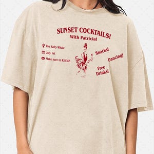 以下が含まれることがあります： 赤いテキストとグラフィックイラストが施されたベージュのTシャツ。テキストには「SUNSET COCKTAILS! With Patricia! The Salty Whale, July 1st, Make sure to R.S.V.P., Snacks!, Dancing!, Free Drinks!」と書かれています。中央のイラストはグラスを持つ人物を描いています。