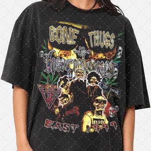 Puede incluir: Camiseta negra con un estampado gráfico del grupo de rap Bone Thugs-n-Harmony. El gráfico presenta el nombre del grupo en letras doradas, una calavera con llamas y las palabras "East 1999".
