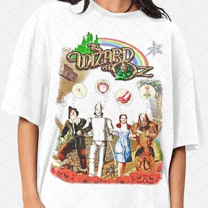 May include: White t-shirt with a graphic print of the characters from the Wizard of Oz. The graphic includes the words "The Wizard of Oz" and images of Dorothy, the Scarecrow, the Tin Man, and the Cowardly Lion. The graphic also includes images of a ruby slipper, a heart, a medal, and a house.