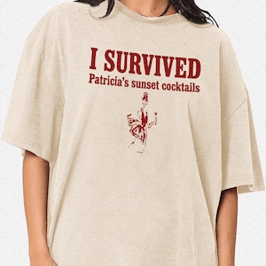 Puede incluir: Una camiseta beige de manga corta con texto en rojo oscuro que dice I SURVIVED Patricia's sunset cocktails. Debajo del texto hay una ilustración gráfica en rojo oscuro de una persona sosteniendo una copa de cóctel, con estilo desgastado.