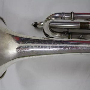 May include: A silver trumpet with an engraved design on the bell. The trumpet has a silver finish and is in good condition.