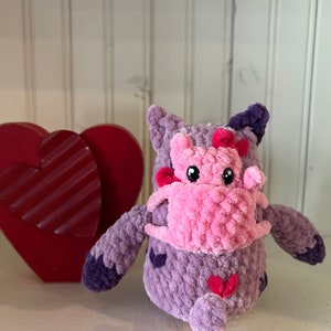May include: A crocheted plush toy in the shape of a purple monster with pink accents. The monster has a pink heart on its chest and a pink heart on its tail. The monster is holding a pink heart in its arms.