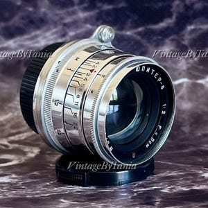 Può includere: Un obiettivo per fotocamera vintage con un design argento e nero. L'obiettivo presenta una serie di numeri e marcature, tra cui "ЮПИТЕР-8" e "1:2 F=5CM". L'obiettivo è posto su una base nera, con il testo "Vintage By Tania" visibile.