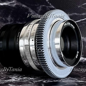 May include: A vintage camera lens with a black body and silver and white detailing. The lens has the text "HELIOS-44M 2/58" and "M42-NEX" printed on it. The lens is set against a dark, textured background.