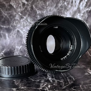 Peut inclure: Un objectif d'appareil photo noir avec une bague argentée et le texte "HELIOS-44M" imprimé dessus. L'objectif est doté d'un bouchon d'objectif noir et d'un pare-soleil noir.